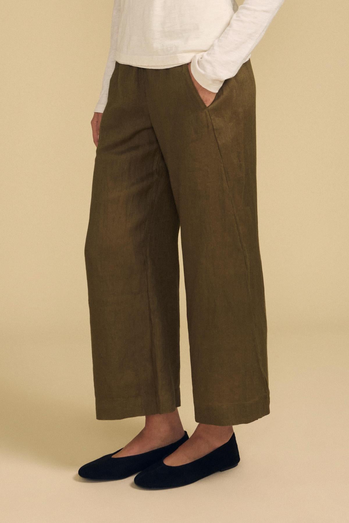 Wearing the Velvet by Graham & Spencer LOLA LINEN PANT in olive green, styled with an elastic waist and wide leg, paired with a long-sleeve white top and black flats. The person stands with hands in pockets against a plain beige background.-42097155735745
