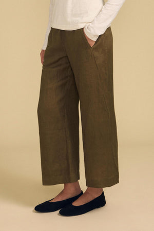 Wearing the Velvet by Graham & Spencer LOLA LINEN PANT in olive green, styled with an elastic waist and wide leg, paired with a long-sleeve white top and black flats. The person stands with hands in pockets against a plain beige background.