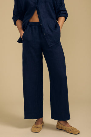 A person stands against a beige background with hands in pockets, wearing the LOLA LINEN PANT by Velvet by Graham & Spencer—loose, wide-leg linen pants with an elastic waist in dark blue—paired with a matching button-up shirt and tan woven slip-on shoes.