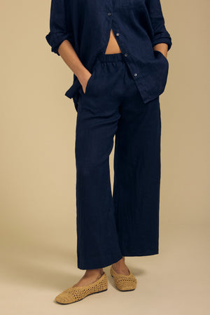 A person wearing the Velvet by Graham & Spencer LOLA LINEN PANT in navy, styled with a matching navy button-up shirt and beige woven slip-on shoes, stands with hands in pockets against a plain beige background.