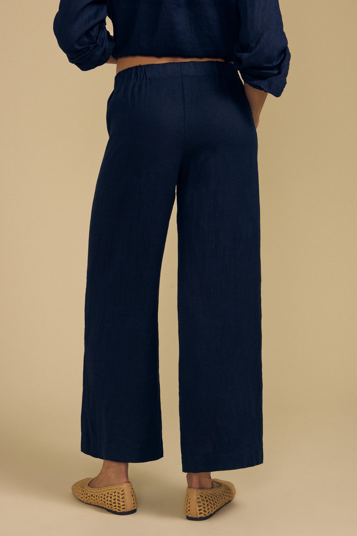 Wearing the Velvet by Graham & Spencer LOLA LINEN PANT in navy—a wide leg linen style with an elastic waist—a person stands facing away, paired with a matching navy top and tan woven slip-on shoes against a beige background.-41960056914113