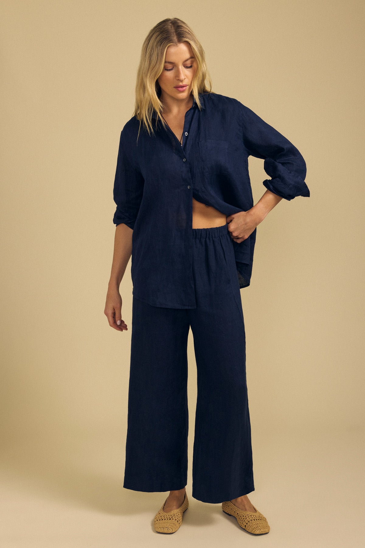 A woman stands against a plain beige background wearing the Velvet by Graham & Spencer LOLA LINEN PANT—a pair of wide-leg, navy linen pants with an elastic waist—styled with a loose-fitting, navy button-up shirt and beige woven slip-on shoes.