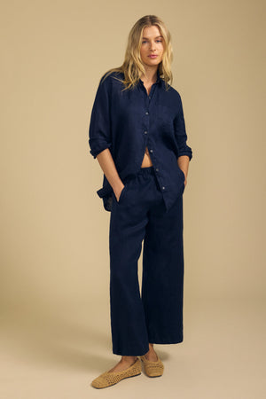 A person stands against a beige background wearing a navy button-up shirt, the LOLA LINEN PANT by Velvet by Graham & Spencer—navy wide leg linen pants with an elastic waist—and tan woven slip-on shoes.