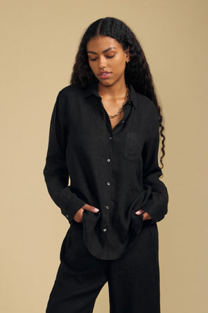 A woman stands against a plain beige background with her hands in her pockets, wearing the loose-fitting, breathable MULHOLLAND LINEN SHIRT and matching pants by Velvet by Jenny Graham.