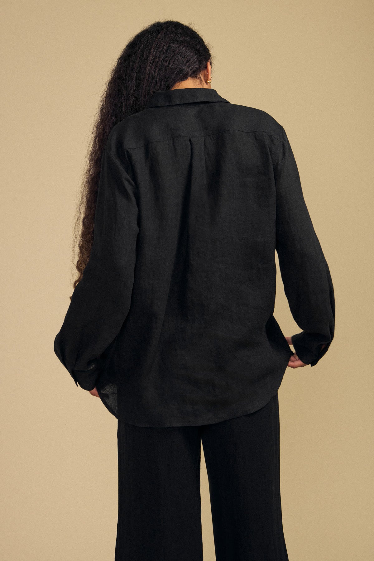 A person with long curly hair is shown from the back against a plain beige background, wearing the MULHOLLAND LINEN SHIRT by Velvet by Jenny Graham in black, styled with black pants.