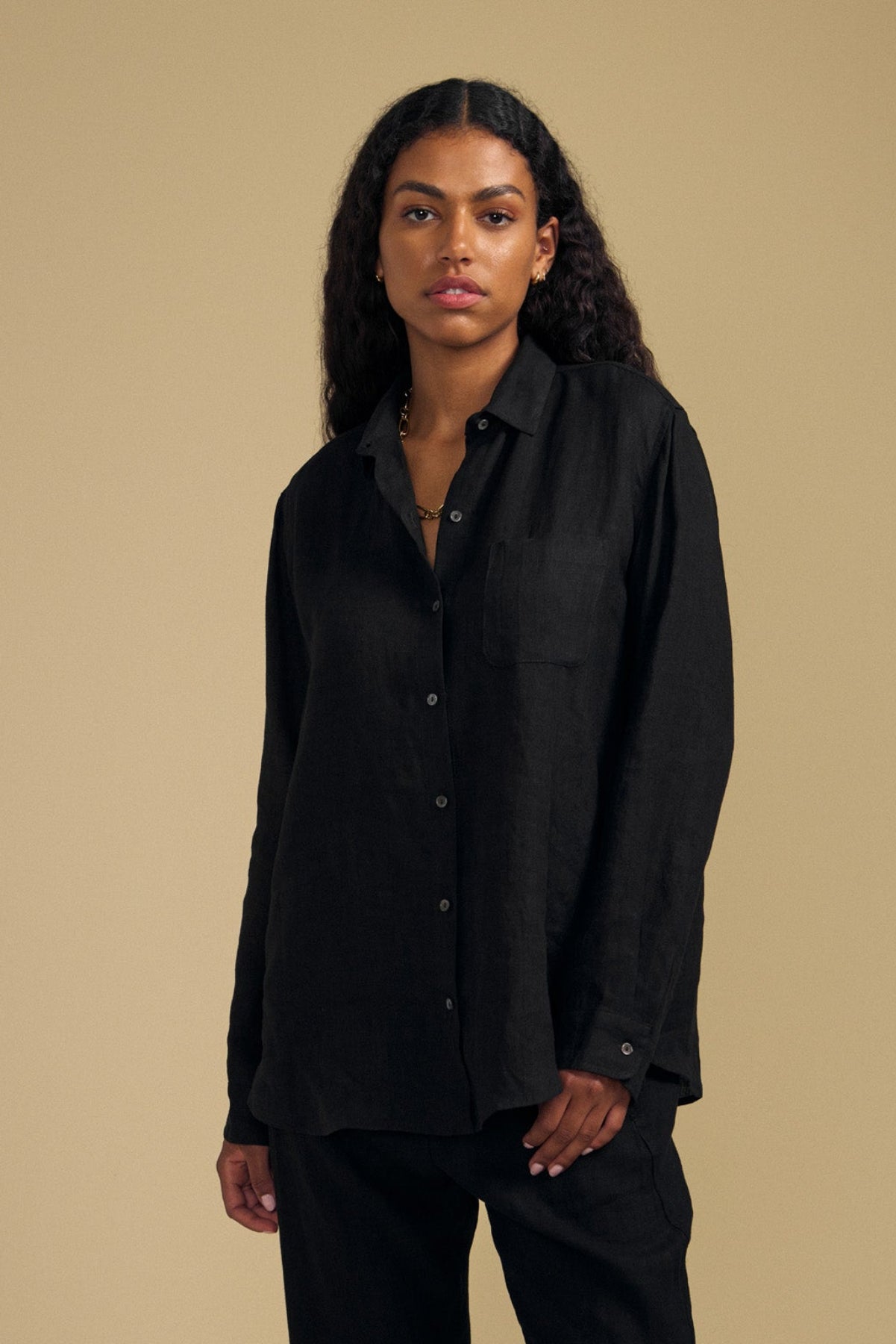 A person with long curly hair wears the MULHOLLAND LINEN SHIRT by Velvet by Jenny Graham in black, styled loosely with matching black pants, standing against a plain beige background.