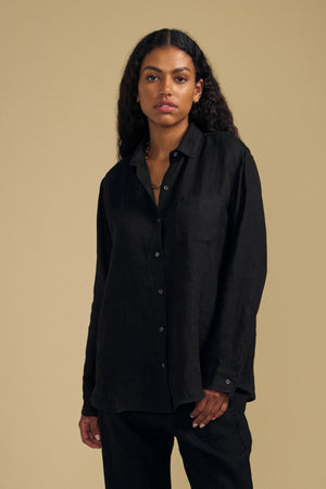 A person with long curly hair wears the MULHOLLAND LINEN SHIRT by Velvet by Jenny Graham in black, styled loosely with matching black pants, standing against a plain beige background.