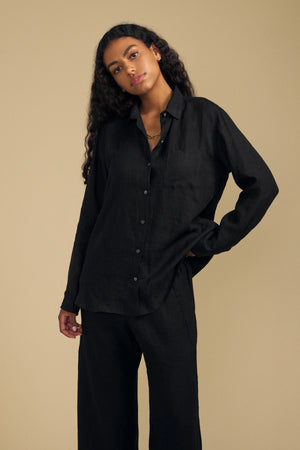 Woman with long curly hair wearing the Velvet by Jenny Graham MULHOLLAND LINEN SHIRT, a loose and lightweight black button-up, paired with matching black pants, standing against a plain beige background.
