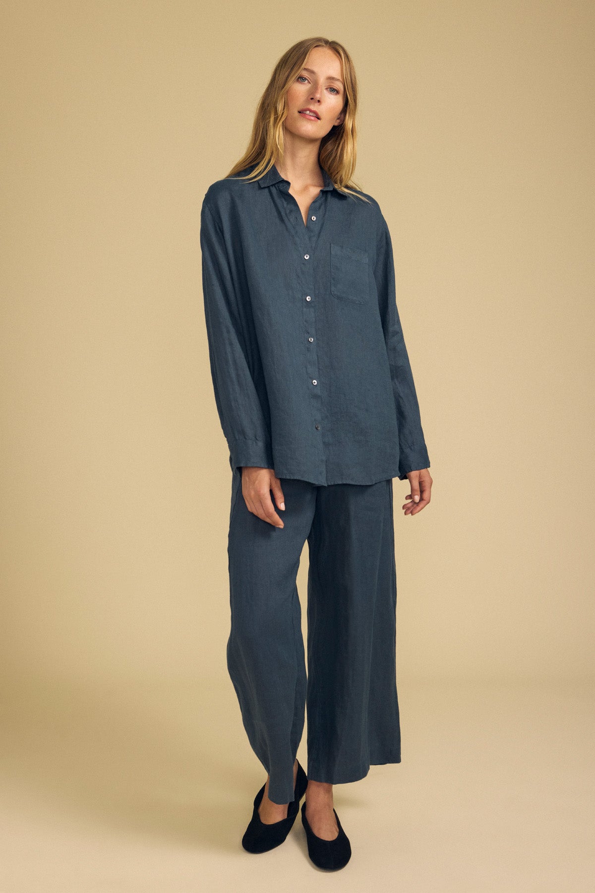 A person stands against a beige background wearing relaxed fit, wide leg LOLA LINEN PANT by Velvet by Graham & Spencer in dark blue, paired with a loose-fitting, long-sleeve button-up shirt.