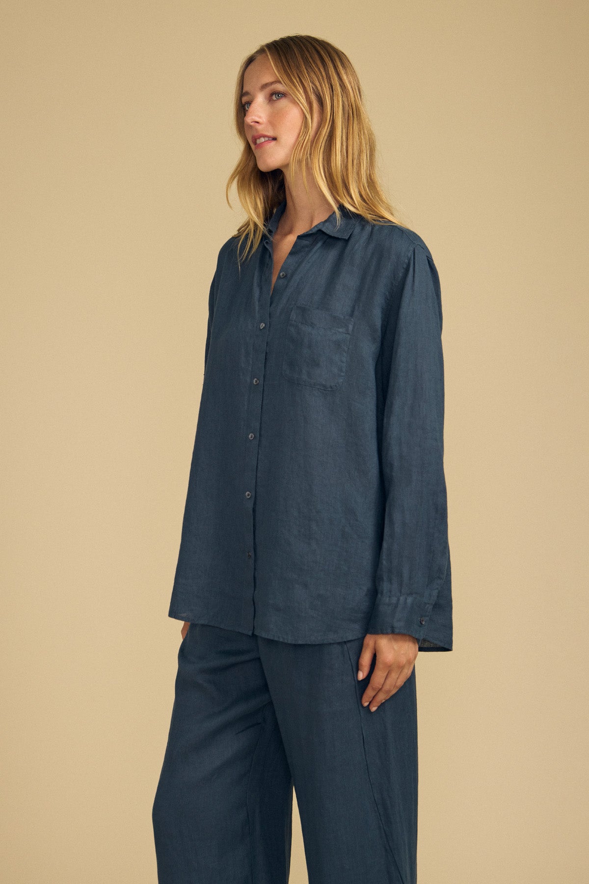 Wearing the MULHOLLAND LINEN SHIRT by Velvet by Jenny Graham, a person with long blonde hair poses against a plain beige background. The shirt is a loose-fitting, dark blue button-up made from breathable linen and is paired with matching pants.