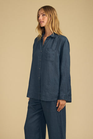 Wearing the MULHOLLAND LINEN SHIRT by Velvet by Jenny Graham, a person with long blonde hair poses against a plain beige background. The shirt is a loose-fitting, dark blue button-up made from breathable linen and is paired with matching pants.