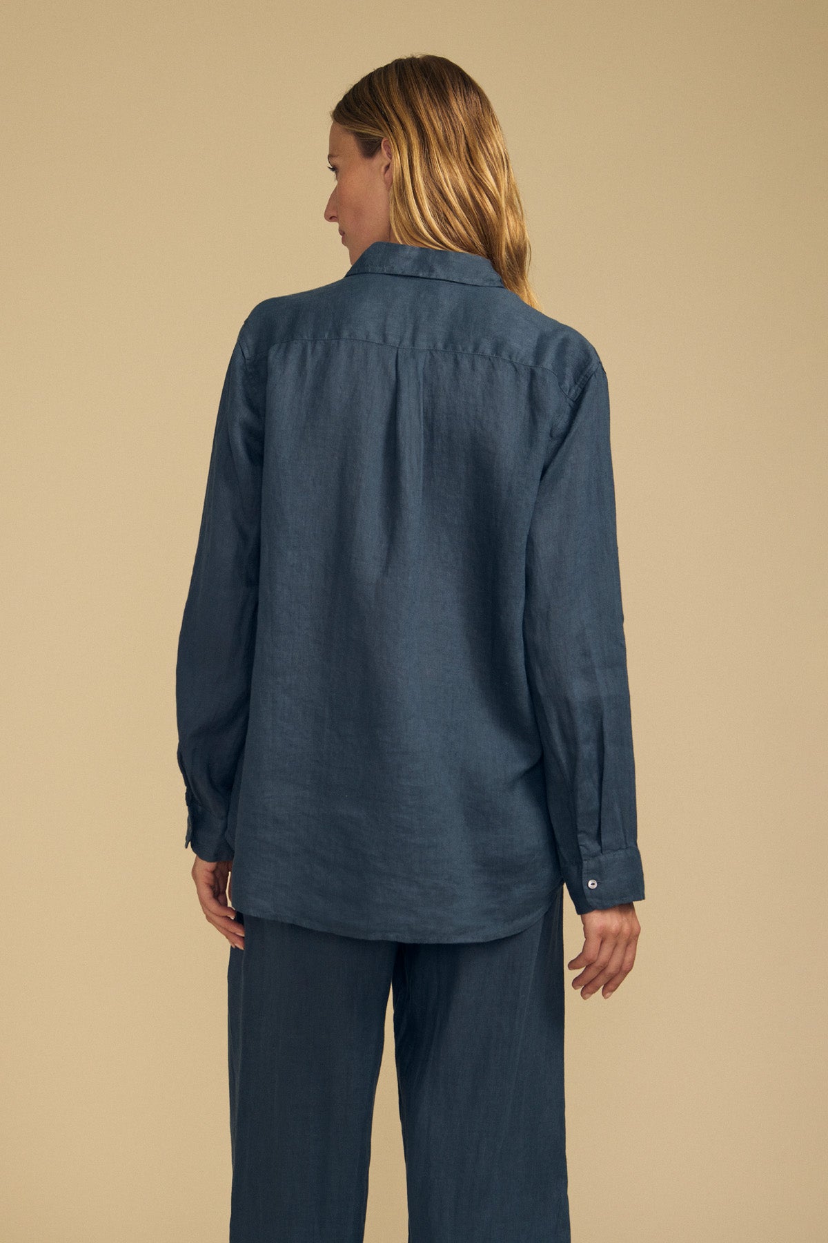A person with long hair is shown from the back against a plain beige background, wearing the MULHOLLAND LINEN SHIRT by Velvet by Jenny Graham—a breathable linen shirt in a relaxed silhouette—paired with matching blue pants.
