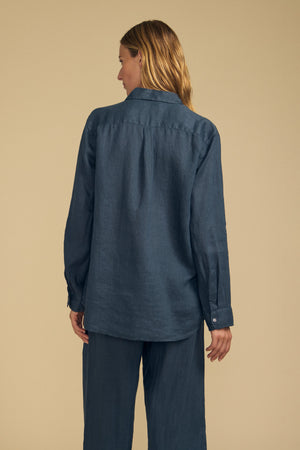 A person with long hair is shown from the back against a plain beige background, wearing the MULHOLLAND LINEN SHIRT by Velvet by Jenny Graham—a breathable linen shirt in a relaxed silhouette—paired with matching blue pants.