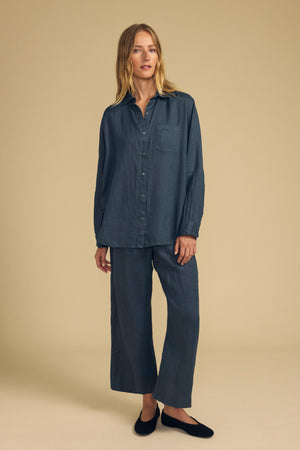 A woman stands against a beige background, dressed in the MULHOLLAND LINEN SHIRT by Velvet by Jenny Graham—a loose-fitting, dark blue button-up paired with matching wide-leg pants and black flat shoes. The relaxed silhouette of her ensemble exudes understated elegance, while the breathable linen fabric ensures all-day comfort.