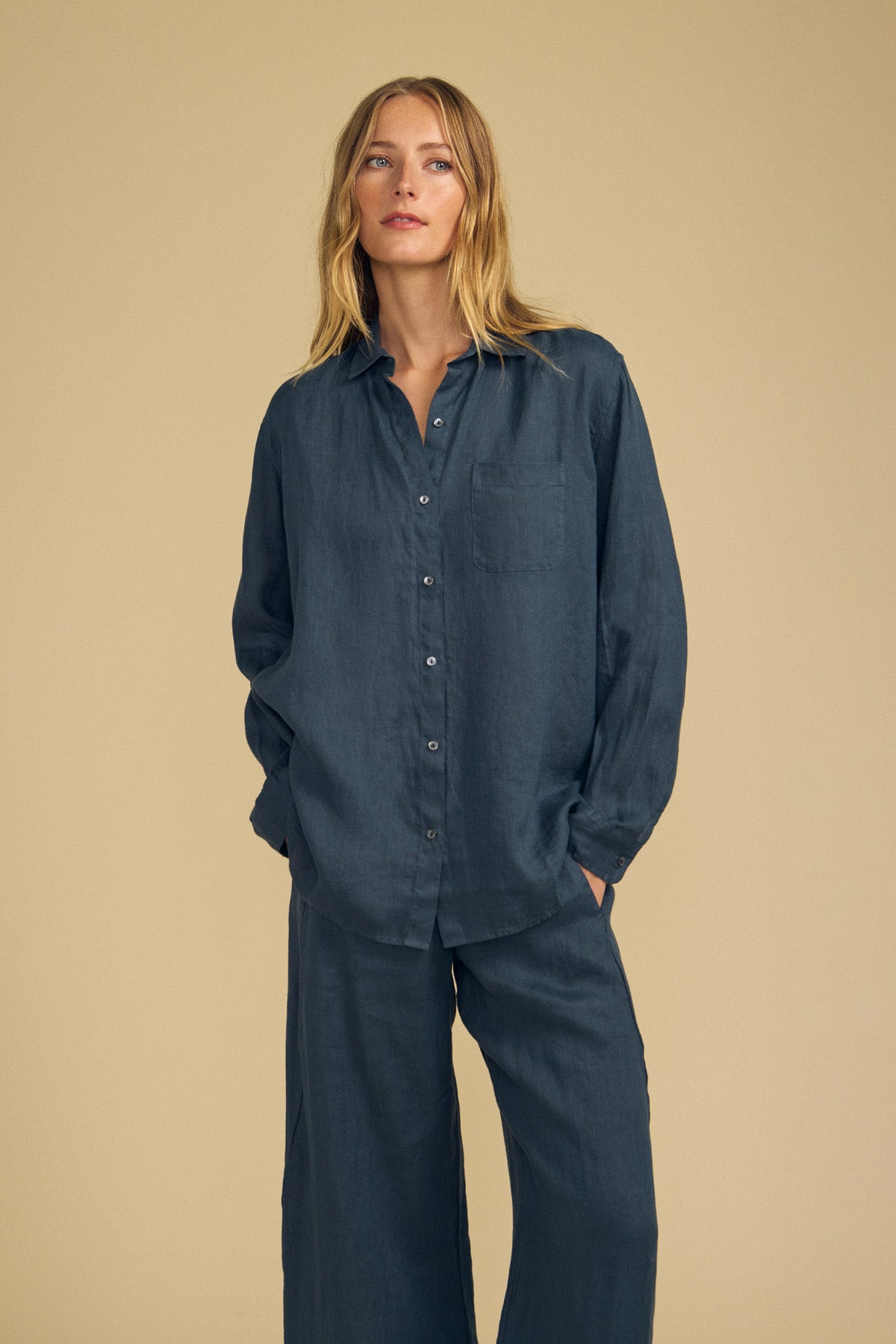 A person stands against a beige background wearing the MULHOLLAND LINEN SHIRT in dark blue by Velvet by Jenny Graham, styled with matching wide-leg pants. The ensemble features a relaxed silhouette, with hands casually placed in pockets.
