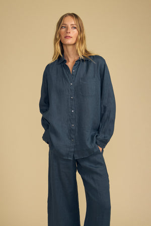 A person stands against a beige background wearing the MULHOLLAND LINEN SHIRT in dark blue by Velvet by Jenny Graham, styled with matching wide-leg pants. The ensemble features a relaxed silhouette, with hands casually placed in pockets.