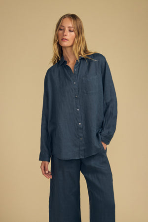 A woman stands against a beige background wearing the MULHOLLAND LINEN SHIRT by Velvet by Jenny Graham, a loose-fitting, long-sleeved blue button-up shirt paired with matching wide-leg pants for a relaxed silhouette.