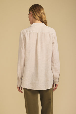 A person with long, light brown hair is shown from the back wearing the MULHOLLAND LINEN SHIRT by Velvet by Jenny Graham in beige, paired with olive green pants, against a plain beige background.
