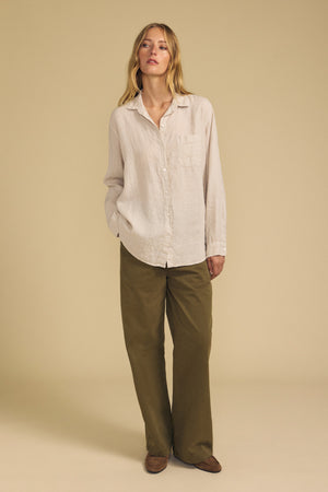 A person stands against a beige background wearing the MULHOLLAND LINEN SHIRT by Velvet by Jenny Graham—a light beige breathable linen button-up—paired with olive green wide-leg pants that create a relaxed silhouette and brown shoes.