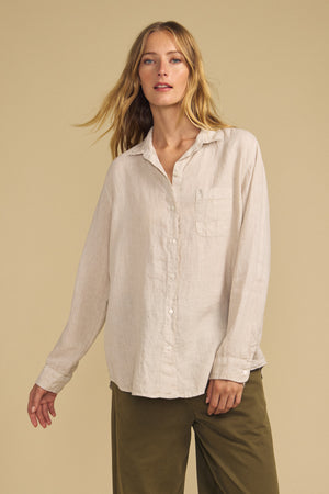 A person wearing the MULHOLLAND LINEN SHIRT by Velvet by Jenny Graham—a relaxed silhouette beige button-up with a chest pocket—paired with olive green pants, stands against a plain beige background.