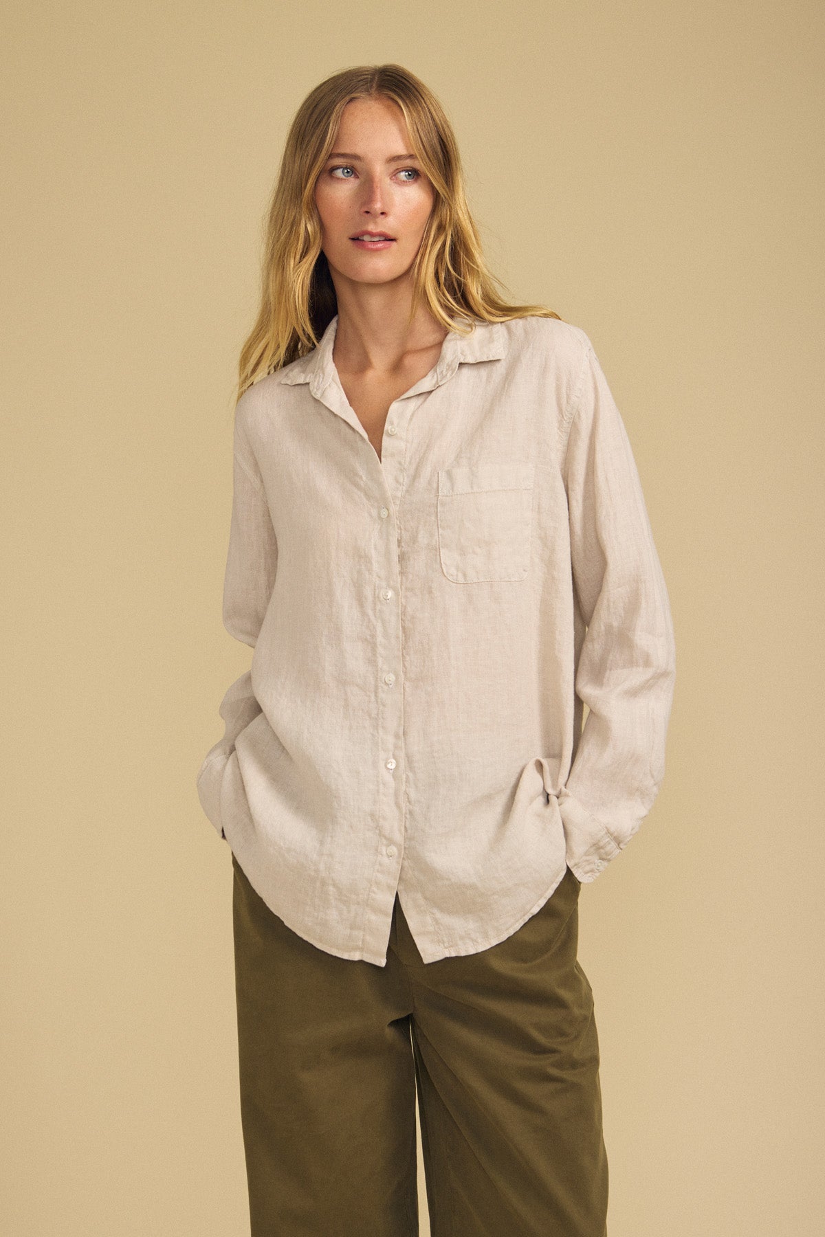 A person wearing the Velvet by Jenny Graham MULHOLLAND LINEN SHIRT in light beige, featuring a breathable linen fabric, button-up front, and chest pocket, paired with olive green pants stands against a plain beige background.