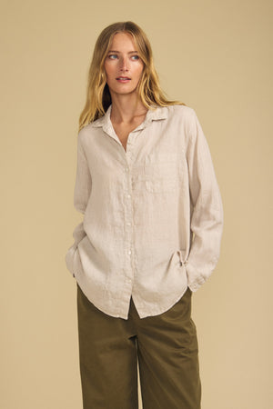 A person wearing the Velvet by Jenny Graham MULHOLLAND LINEN SHIRT in light beige, featuring a breathable linen fabric, button-up front, and chest pocket, paired with olive green pants stands against a plain beige background.