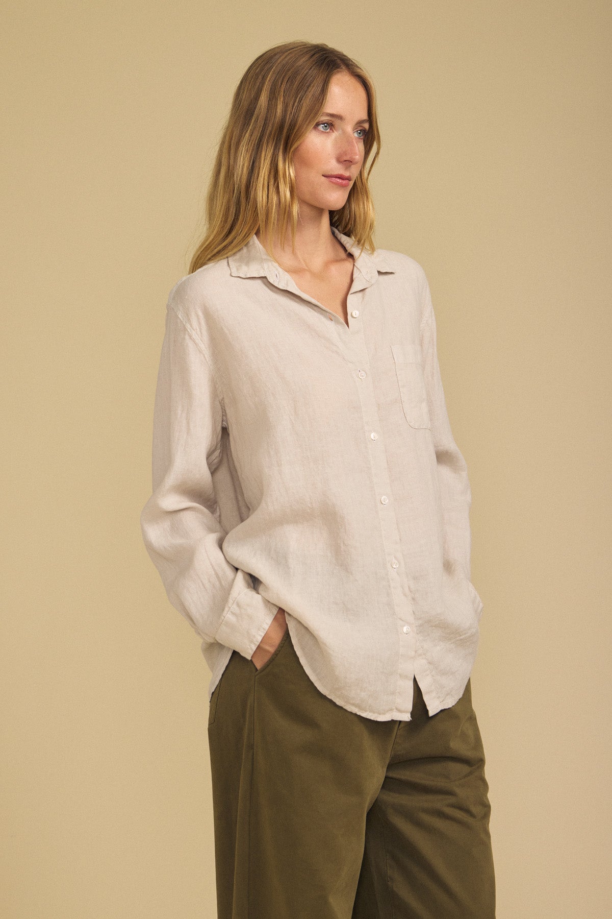 Wearing the MULHOLLAND LINEN SHIRT from Velvet by Jenny Graham in light beige, paired with olive green pants, a person stands with hands in pockets against a plain beige background, highlighting the shirt’s breathable fabric and relaxed silhouette.