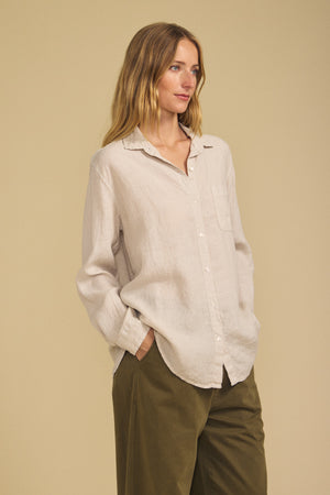Wearing the MULHOLLAND LINEN SHIRT from Velvet by Jenny Graham in light beige, paired with olive green pants, a person stands with hands in pockets against a plain beige background, highlighting the shirt’s breathable fabric and relaxed silhouette.