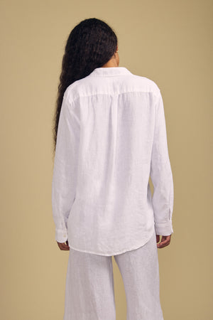 A person stands with their back to the camera, wearing Velvet by Jenny Graham’s MULHOLLAND LINEN SHIRT—a white long-sleeve button-up—paired with matching white pants against a beige background. The relaxed silhouette and airy linen design of the shirt create an effortlessly stylish look.