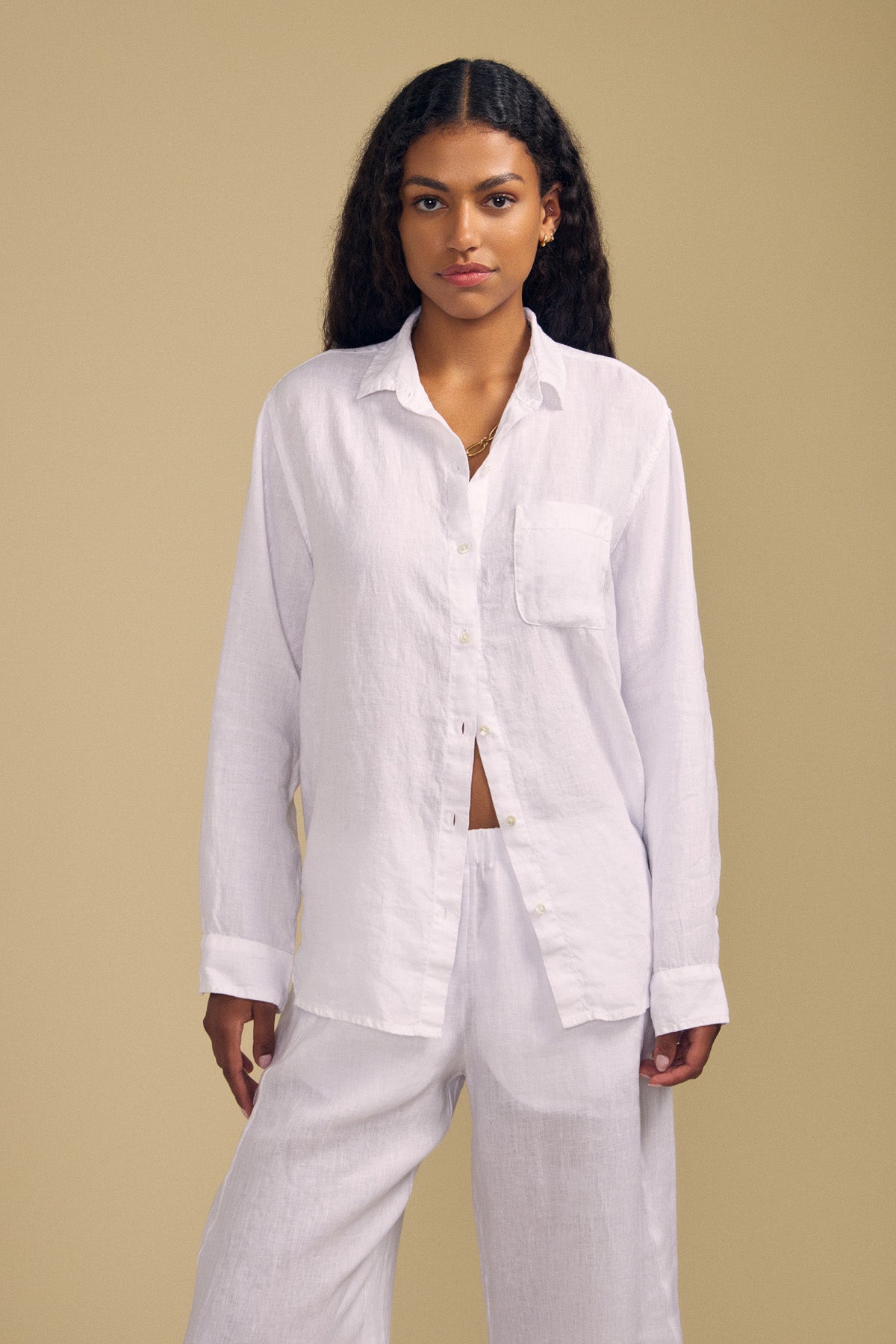 A woman stands against a beige background, facing forward with a neutral expression, wearing the Velvet by Jenny Graham MULHOLLAND LINEN SHIRT in white paired with matching white pants.