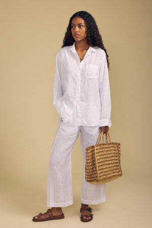 Against a beige background, a woman wears a white linen shirt and pants with brown sandals, carrying the chic MELODY BAG by Velvet by Graham & Spencer.