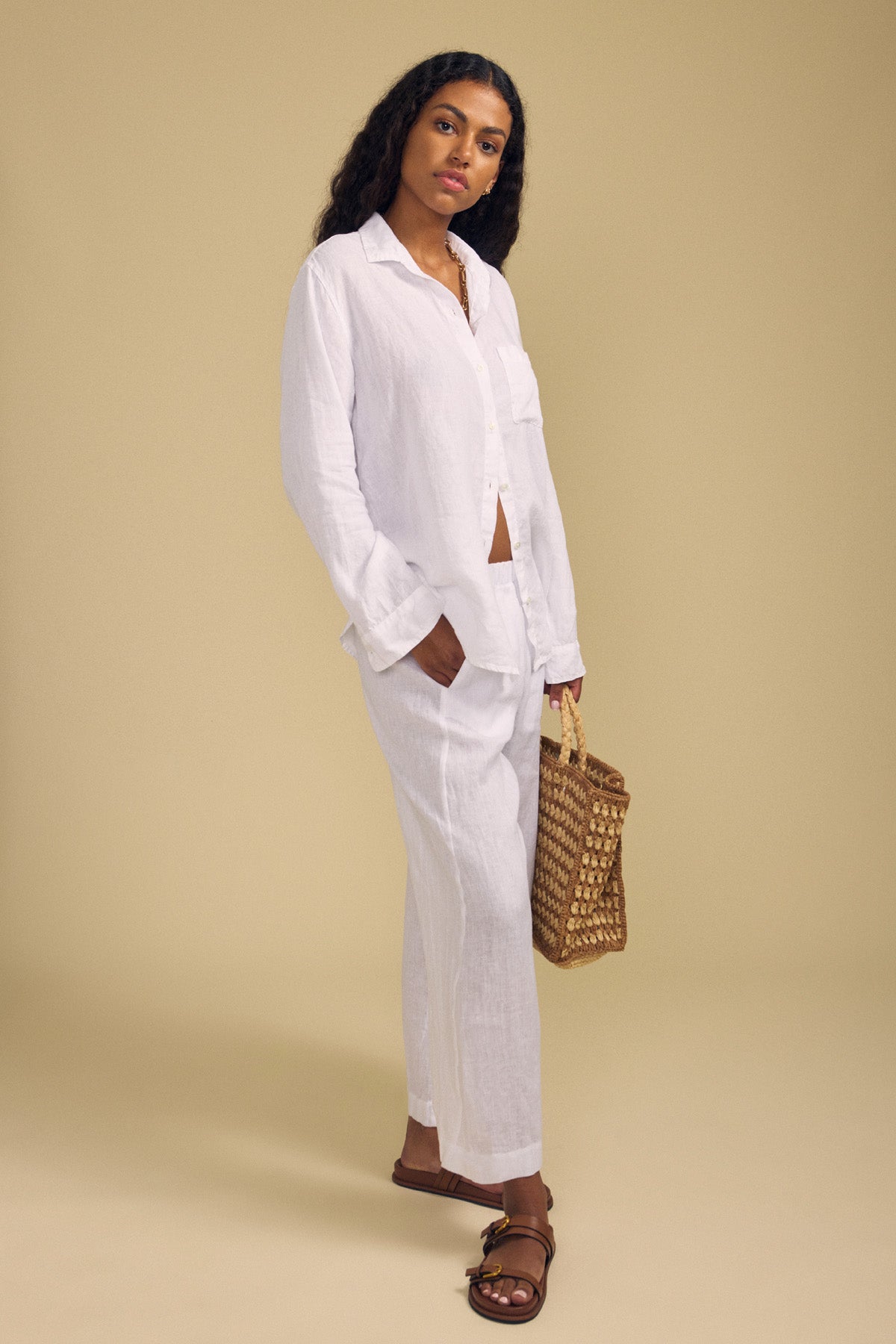 A woman stands against a beige background, wearing the Velvet by Jenny Graham MULHOLLAND LINEN SHIRT and matching pants, paired with brown sandals and holding a woven bag.