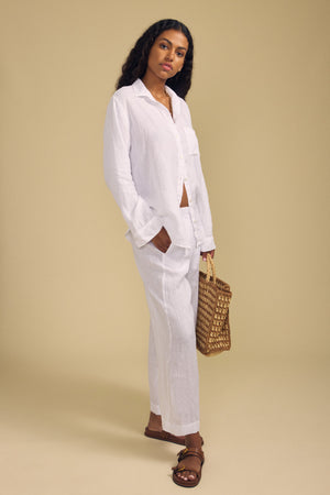 A woman stands against a beige background, wearing the Velvet by Jenny Graham MULHOLLAND LINEN SHIRT and matching pants, paired with brown sandals and holding a woven bag.