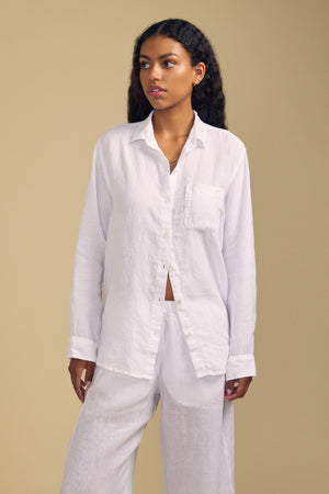 Styled in the MULHOLLAND LINEN SHIRT by Velvet by Jenny Graham and matching white pants, this look features a relaxed silhouette that enhances its effortless appeal against a plain beige background.