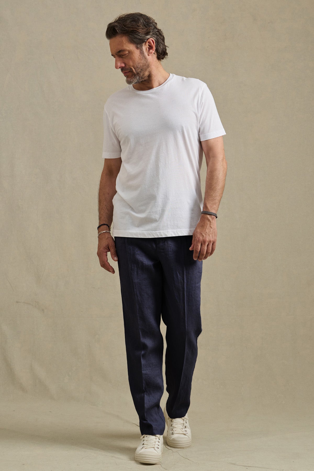 A man in a plain white t-shirt, Velvet by Graham & Spencer ELLIOTT PANT, and white sneakers stands against a neutral background, gazing down and to the side.-41435144028353
