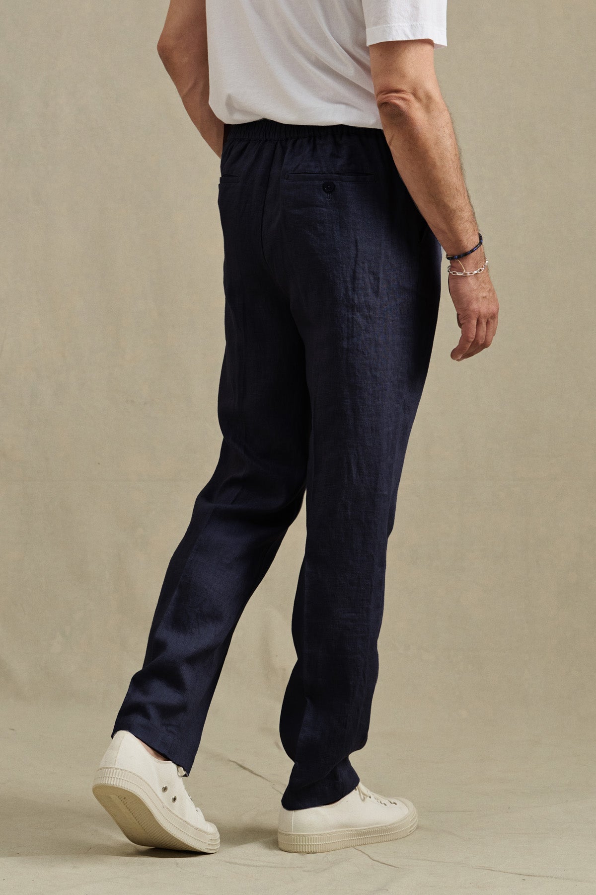 A person is standing on a neutral background, facing away, wearing a white t-shirt, the ELLIOTT PANT in dark blue by Velvet by Graham & Spencer, and white sneakers.