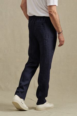 A person is standing on a neutral background, facing away, wearing a white t-shirt, the ELLIOTT PANT in dark blue by Velvet by Graham & Spencer, and white sneakers.