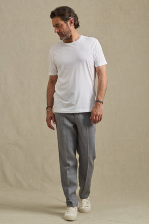 A man stands on a plain beige background, looking down with his hands relaxed at his sides, wearing a white t-shirt, Velvet by Graham & Spencer ELLIOTT PANT slim-fit linen trousers, and white sneakers.