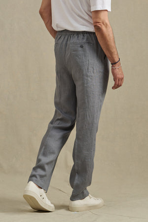 A person wearing the ELLIOTT PANT by Velvet by Graham & Spencer, a white t-shirt, and white sneakers stands on a light-colored surface, shown from the back.