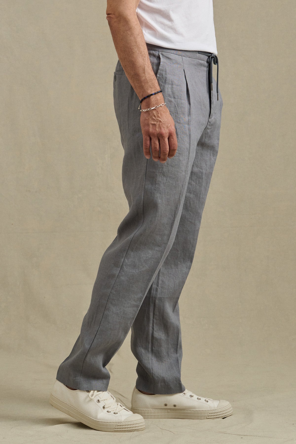 A person wearing the ELLIOTT PANT by Velvet by Graham & Spencer—slim-fit linen trousers with an elastic waist—paired with a white t-shirt, white sneakers, and a bracelet, stands against a neutral background.