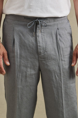 A person shown from waist to mid-thigh wearing the ELLIOTT PANT by Velvet by Graham & Spencer in light gray slim-fit linen, paired with a white shirt.