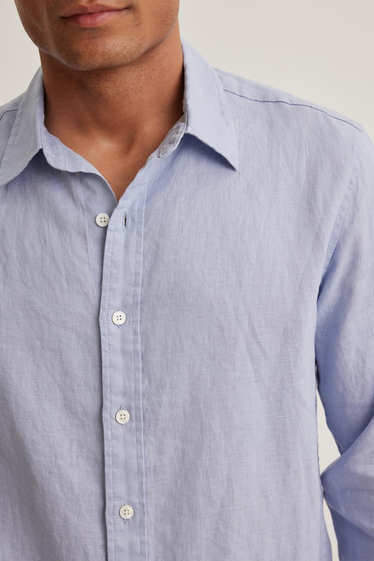 A person wearing a Velvet by Graham & Spencer CARUSO LINEN BUTTON-UP SHIRT stands against a neutral background. The shirt is light blue with a classic collar and white buttons.-38643242369217