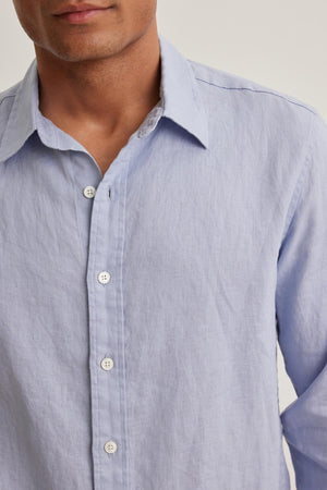 A person wearing a Velvet by Graham & Spencer CARUSO LINEN BUTTON-UP SHIRT stands against a neutral background. The shirt is light blue with a classic collar and white buttons.