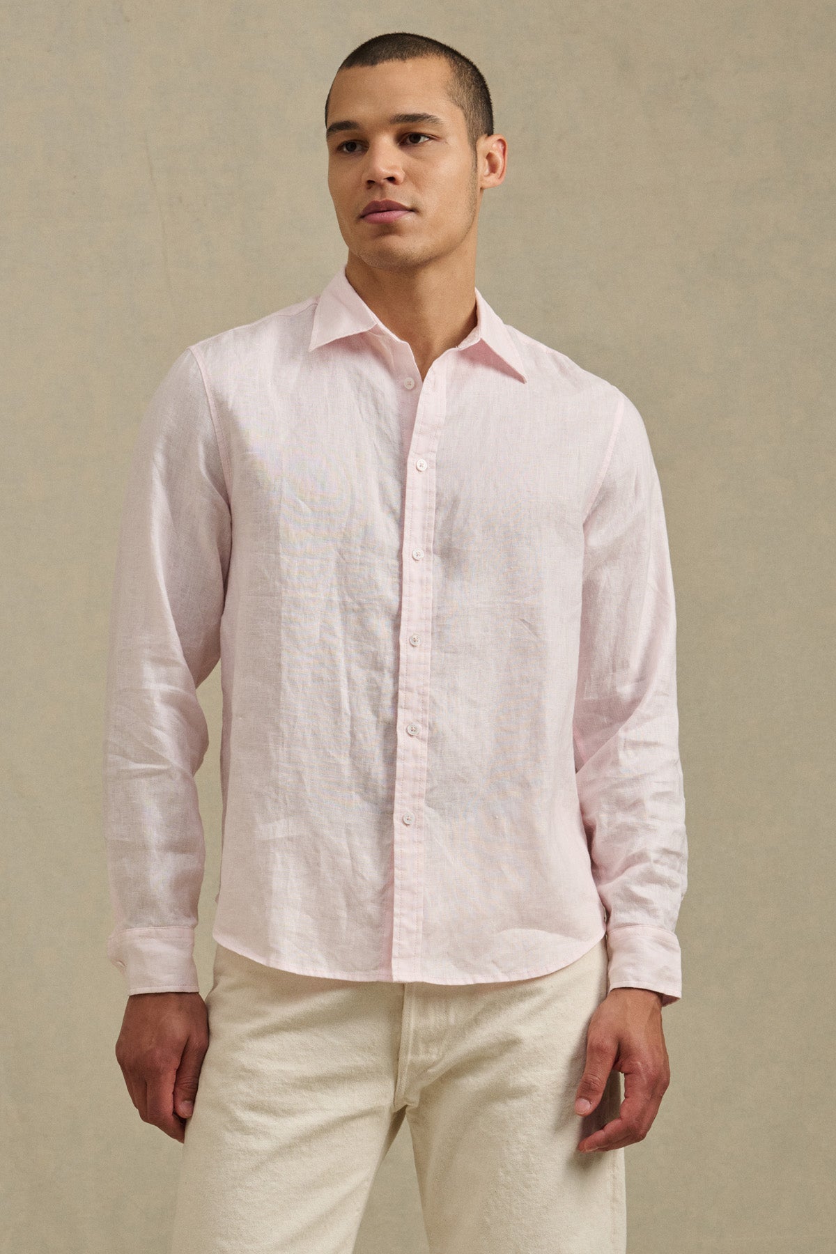 A man wears the CARUSO LINEN SHIRT by Velvet by Graham & Spencer in light pink with off-white pants, standing against a beige background—showcasing versatile, effortless style for any occasion.