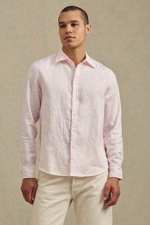 A man wears the CARUSO LINEN SHIRT by Velvet by Graham & Spencer in light pink with off-white pants, standing against a beige background—showcasing versatile, effortless style for any occasion.