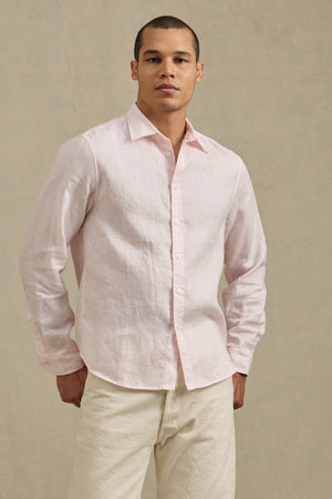 A man stands against a neutral background, wearing the CARUSO LINEN SHIRT by Velvet by Graham & Spencer in light pink with off-white pants—ideal for versatile styling and effortless layering.