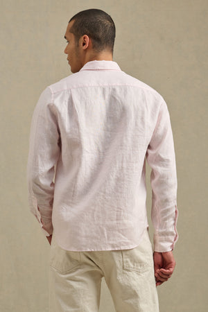 A person faces away, wearing the CARUSO LINEN SHIRT by Velvet by Graham & Spencer in light pink with off-white pants against a neutral background—a versatile piece for easy layering.