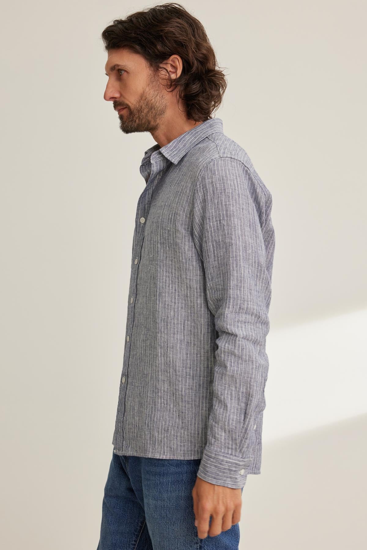   A man with long hair and a beard stands in profile, wearing a CARUSO LINEN BUTTON-UP SHIRT by Velvet by Graham & Spencer, paired with blue jeans against a plain background. 