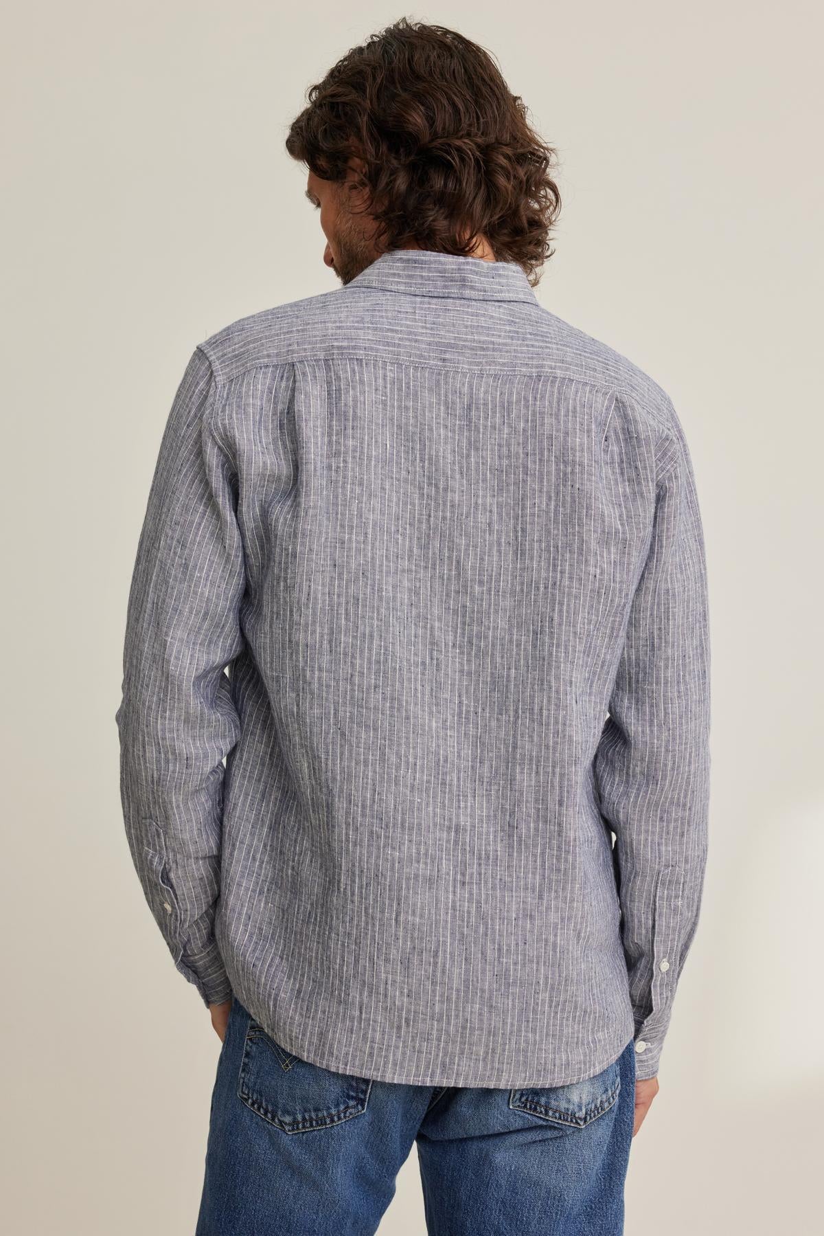   A person with wavy hair is wearing a CARUSO LINEN BUTTON-UP SHIRT by Velvet by Graham & Spencer, paired with blue jeans, facing away. 