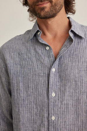 A man with medium-length hair and a beard wears a partially unbuttoned CARUSO LINEN BUTTON-UP SHIRT by Velvet by Graham & Spencer in blue pinstripe, set against a plain background.